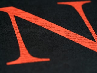 A large red letter N is overlaid on a black background, with small typed text visible within the letter. The text provides a textured appearance, and the color contrast is striking.