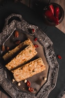 A decorative silver tray holds three rectangular pieces of baked pastry or bread with a textured, crunchy outer layer. Surrounding the pastries are a few red chilies, pecans, and scattered peanuts. A dark cloth underlies the tray, and near the edge, a bowl of vibrant red chili peppers adds a pop of color.