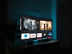 A television screen displaying a user interface with popular streaming service apps including Netflix, Sky, Apple TV, Prime Video, and others. The screen also shows images from movies or shows above the apps. The room is dimly lit with a potted plant visible on the side.