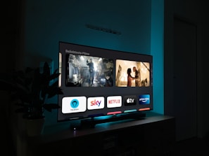 A television screen displaying a user interface with popular streaming service apps including Netflix, Sky, Apple TV, Prime Video, and others. The screen also shows images from movies or shows above the apps. The room is dimly lit with a potted plant visible on the side.