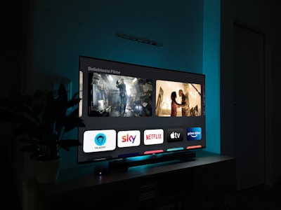 A television screen displaying a user interface with popular streaming service apps including Netflix, Sky, Apple TV, Prime Video, and others. The screen also shows images from movies or shows above the apps. The room is dimly lit with a potted plant visible on the side.