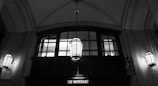An elegant, arched interior space characterized by large, detailed hanging lanterns. The ceiling is vaulted with a geometric design, and a row of windows with a classic frame is visible against the wall. Below the windows, a sign reads 'The Merchant,' and the area is lit with soft, ambient lighting.