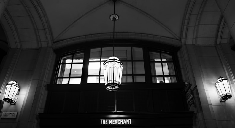 An elegant, arched interior space characterized by large, detailed hanging lanterns. The ceiling is vaulted with a geometric design, and a row of windows with a classic frame is visible against the wall. Below the windows, a sign reads 'The Merchant,' and the area is lit with soft, ambient lighting.