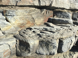 Layers of sedimentary rock formation with visible strata patterns. The texture appears rugged and weathered, showing shades of gray, brown, and hints of rust. The geological structure suggests erosion and time-worn surfaces.