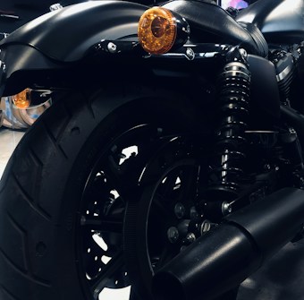 A close-up view of a motorcycle's rear section featuring a large black tire, a metallic suspension coil, and an exhaust pipe. The orange indicators add a subtle contrast to the predominantly black and metallic components. The overall image conveys a sleek and polished appearance with attention to detail in the mechanical parts.