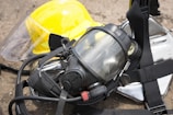 A close-up of a protective gas mask with straps and a transparent visor placed on top of a reflective metallic surface, next to a bright yellow safety helmet with a face shield attached.