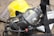 A close-up of a protective gas mask with straps and a transparent visor placed on top of a reflective metallic surface, next to a bright yellow safety helmet with a face shield attached.