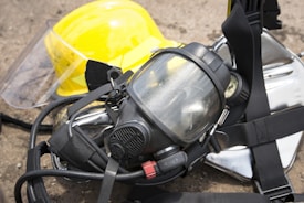 A close-up of a protective gas mask with straps and a transparent visor placed on top of a reflective metallic surface, next to a bright yellow safety helmet with a face shield attached.