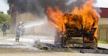 A wooden structure engulfed in bright orange flames with thick black smoke billowing upwards. Firefighters are present, wearing protective gear including helmets, and actively using a hose to spray water on the fire. The background includes a brick building and grassy area.