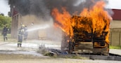 A wooden structure engulfed in bright orange flames with thick black smoke billowing upwards. Firefighters are present, wearing protective gear including helmets, and actively using a hose to spray water on the fire. The background includes a brick building and grassy area.