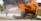 A wooden structure engulfed in bright orange flames with thick black smoke billowing upwards. Firefighters are present, wearing protective gear including helmets, and actively using a hose to spray water on the fire. The background includes a brick building and grassy area.