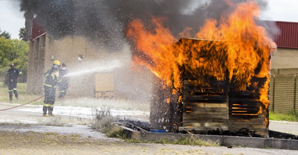 A wooden structure engulfed in bright orange flames with thick black smoke billowing upwards. Firefighters are present, wearing protective gear including helmets, and actively using a hose to spray water on the fire. The background includes a brick building and grassy area.