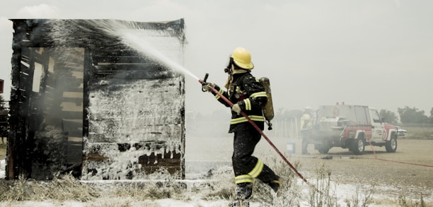 A firefighter in full protective gear is actively spraying water on a burning wooden structure, which is covered in white foam. A fire truck is parked in the background, with a couple of firefighters nearby, indicating an emergency response scene. The environment appears to be an open, rural area under a cloudy sky.