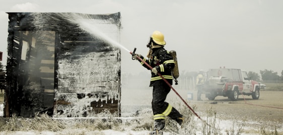 A firefighter in full protective gear is actively spraying water on a burning wooden structure, which is covered in white foam. A fire truck is parked in the background, with a couple of firefighters nearby, indicating an emergency response scene. The environment appears to be an open, rural area under a cloudy sky.