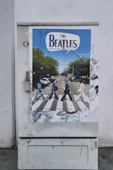 A poster is displayed on an outdoor utility box, featuring a comic-style depiction of a famous crosswalk scene with four figures walking across. The background includes trees, vehicles, and buildings. The top of the poster has bold text that appears to be a title or heading.