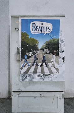 A poster is displayed on an outdoor utility box, featuring a comic-style depiction of a famous crosswalk scene with four figures walking across. The background includes trees, vehicles, and buildings. The top of the poster has bold text that appears to be a title or heading.