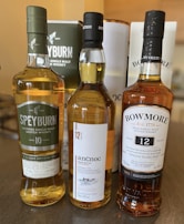 Three bottles of Scotch whisky are positioned side by side on a countertop. The labels identify them as Speyburn 10-year-old, anCnoc 12-year-old, and Bowmore 12-year-old. Each bottle has a distinct label with varying designs and colors.