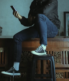 A person sitting casually on a wooden surface with one leg crossed over the other, holding a smartphone. The individual is wearing a black jacket, dark blue jeans, and black canvas sneakers with white soles. A black stool supports the person's feet. The setting seems to resemble an indoor space with detailed wood carvings on the furniture.