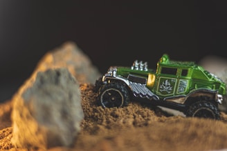 A small green toy truck navigating through a sandy terrain with large rocks in the foreground. The truck has detailed designs and insignia on the side, with a shiny metallic exterior, suggesting it is a model vehicle.