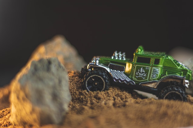 A small green toy truck navigating through a sandy terrain with large rocks in the foreground. The truck has detailed designs and insignia on the side, with a shiny metallic exterior, suggesting it is a model vehicle.