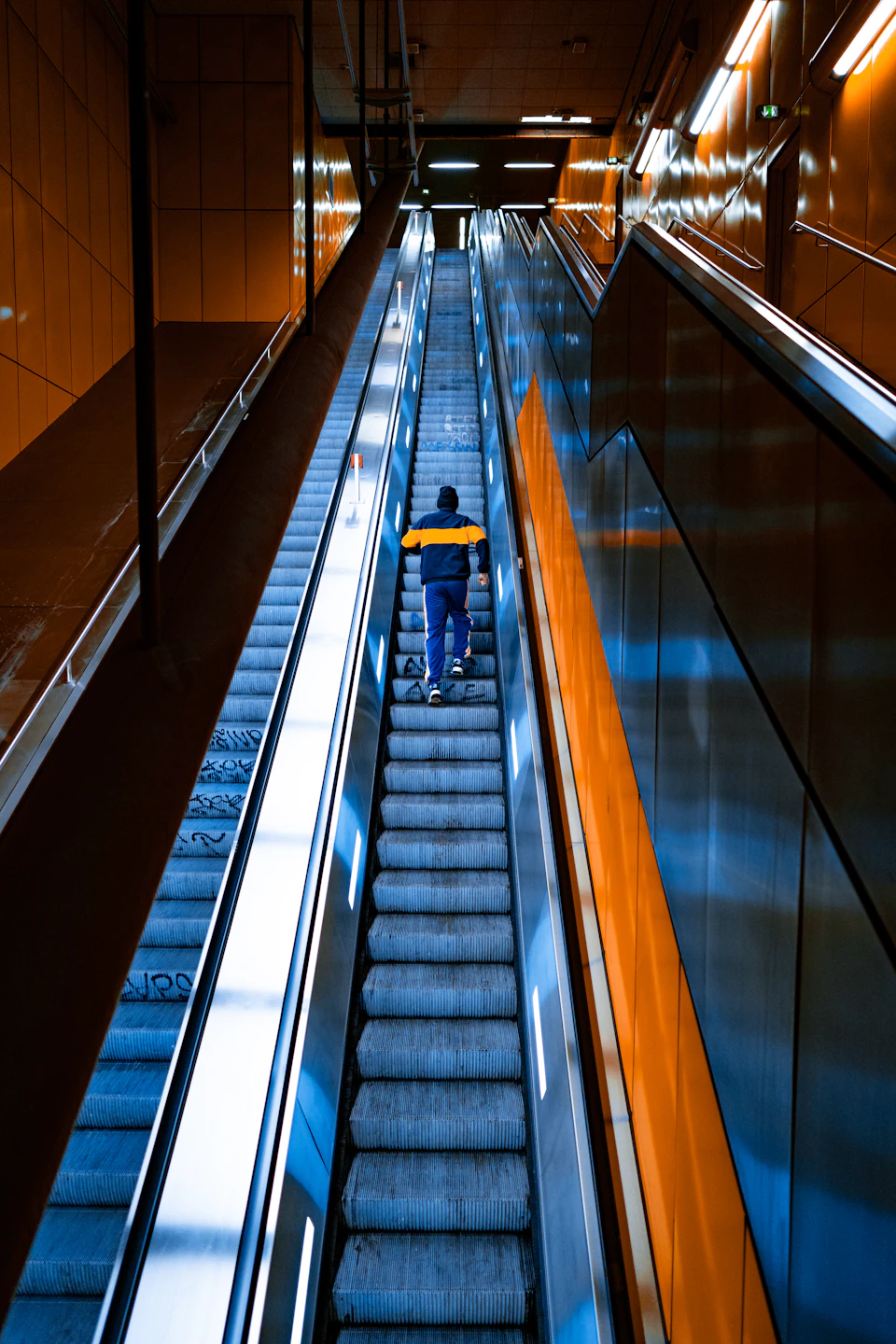 Person ascending escalator in subway station with blue jacket and yellow striped pants