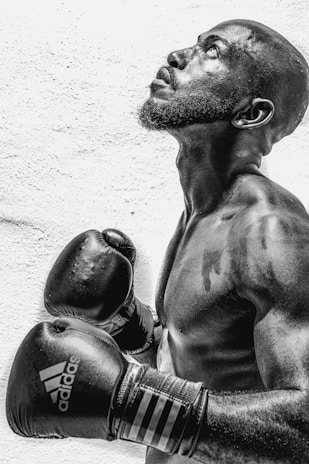 A black and white photograph of a muscular man looking upwards, wearing boxing gloves branded with a prominent logo. His stance is focused and poised, with visible perspiration indicating exertion.