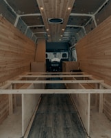 The interior of a van is under construction, featuring wooden paneling along the walls and ceiling. The floor has a dark wood finish, and the framework for what appears to be cabinetry or furniture is visible. Recessed lighting is installed in the ceiling.