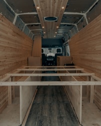 The interior of a van is under construction, featuring wooden paneling along the walls and ceiling. The floor has a dark wood finish, and the framework for what appears to be cabinetry or furniture is visible. Recessed lighting is installed in the ceiling.