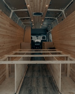 The interior of a van is under construction, featuring wooden paneling along the walls and ceiling. The floor has a dark wood finish, and the framework for what appears to be cabinetry or furniture is visible. Recessed lighting is installed in the ceiling.