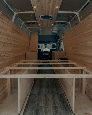 The interior of a van is under construction, featuring wooden paneling along the walls and ceiling. The floor has a dark wood finish, and the framework for what appears to be cabinetry or furniture is visible. Recessed lighting is installed in the ceiling.