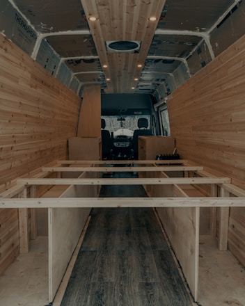 The interior of a van is under construction, featuring wooden paneling along the walls and ceiling. The floor has a dark wood finish, and the framework for what appears to be cabinetry or furniture is visible. Recessed lighting is installed in the ceiling.