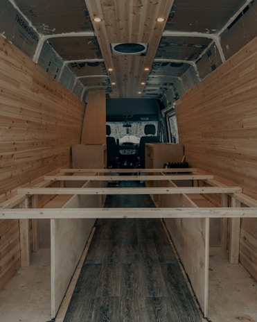 The interior of a van is under construction, featuring wooden paneling along the walls and ceiling. The floor has a dark wood finish, and the framework for what appears to be cabinetry or furniture is visible. Recessed lighting is installed in the ceiling.