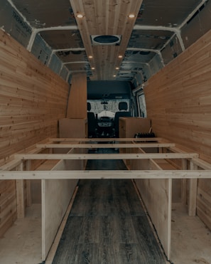The interior of a van is under construction, featuring wooden paneling along the walls and ceiling. The floor has a dark wood finish, and the framework for what appears to be cabinetry or furniture is visible. Recessed lighting is installed in the ceiling.