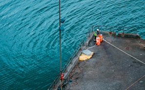 A worker wearing a high-visibility jacket and helmet stands on a dock next to orange buoys and coiled ropes. The person appears to be engaged in work-related activity, possibly related to the maintenance or operation of the dock facilities. The water around the dock is a vibrant blue-green, with gentle ripples.