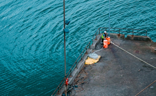 A worker wearing a high-visibility jacket and helmet stands on a dock next to orange buoys and coiled ropes. The person appears to be engaged in work-related activity, possibly related to the maintenance or operation of the dock facilities. The water around the dock is a vibrant blue-green, with gentle ripples.