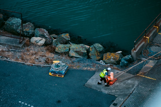 A person wearing a high-visibility vest and hard hat is handling a rope on a waterfront dock. The area is bordered by large, weathered rocks and the water is visible beyond. A coil of rope is resting on a pallet nearby.