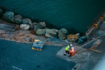 A person wearing a high-visibility vest and hard hat is handling a rope on a waterfront dock. The area is bordered by large, weathered rocks and the water is visible beyond. A coil of rope is resting on a pallet nearby.