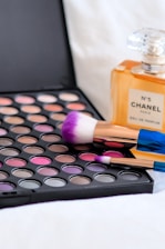 A variety of colorful eyeshadows in a palette accompanied by makeup brushes with purple bristles. A bottle of Chanel No. 5 perfume stands nearby, creating a luxurious and elegant atmosphere.