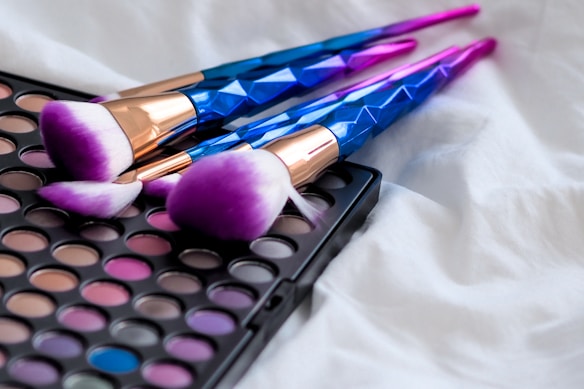 Three makeup brushes with purple-tipped bristles and metallic blue handles are placed on a colorful eyeshadow palette, lying on a white, fluffy fabric.