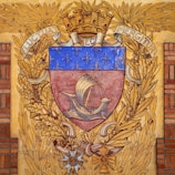 A heraldic shield features a medieval ship with sails, surrounded by a decorative border of golden oak leaves and a laurel wreath. The upper part of the shield is adorned with a series of fleur-de-lis on a blue background. The crest is topped with a mural crown and flanked by the Latin motto 'Fluctuat Nec Mergitur'. Below the shield, there are ornate insignias and decorations.