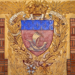 A heraldic shield features a medieval ship with sails, surrounded by a decorative border of golden oak leaves and a laurel wreath. The upper part of the shield is adorned with a series of fleur-de-lis on a blue background. The crest is topped with a mural crown and flanked by the Latin motto 'Fluctuat Nec Mergitur'. Below the shield, there are ornate insignias and decorations.