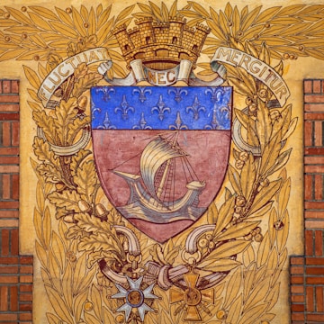 A heraldic shield features a medieval ship with sails, surrounded by a decorative border of golden oak leaves and a laurel wreath. The upper part of the shield is adorned with a series of fleur-de-lis on a blue background. The crest is topped with a mural crown and flanked by the Latin motto 'Fluctuat Nec Mergitur'. Below the shield, there are ornate insignias and decorations.