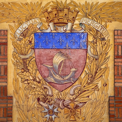 A heraldic shield features a medieval ship with sails, surrounded by a decorative border of golden oak leaves and a laurel wreath. The upper part of the shield is adorned with a series of fleur-de-lis on a blue background. The crest is topped with a mural crown and flanked by the Latin motto 'Fluctuat Nec Mergitur'. Below the shield, there are ornate insignias and decorations.