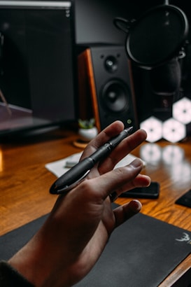 A hand holding a pen, situated in a workspace with a wooden desk. In the background, there is a computer monitor, studio speakers, a microphone, and a smartphone. The scene appears to be a home office or recording studio setup.