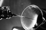 A close-up view of a liquid being poured from a bottle into a glass container. The image is in black and white, capturing the smooth flow of the liquid under ambient lighting. The glass container has a slightly reflective surface, and the background is blurred, emphasizing the fluid motion.