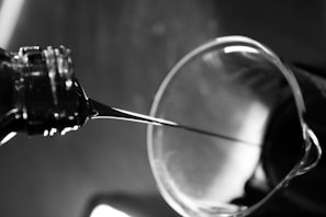 A close-up view of a liquid being poured from a bottle into a glass container. The image is in black and white, capturing the smooth flow of the liquid under ambient lighting. The glass container has a slightly reflective surface, and the background is blurred, emphasizing the fluid motion.