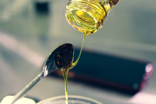 Pouring a stream of golden liquid, likely honey or syrup, into a metal spoon. The liquid is viscous and glistening under the light, creating an elegant and smooth curve as it flows.