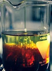 A glass beaker filled with a yellow liquid. The lower part of the liquid is mixed with a swirling, vivid red substance, creating a dynamic and intriguing visual effect within the container. Light reflects off the surface, enhancing the colors and creating a sense of motion.