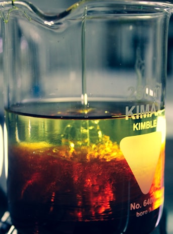 A glass beaker filled with a yellow liquid. The lower part of the liquid is mixed with a swirling, vivid red substance, creating a dynamic and intriguing visual effect within the container. Light reflects off the surface, enhancing the colors and creating a sense of motion.