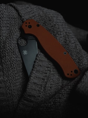 A folding knife with an orange handle partially concealed between layers of a dark textured fabric. The blade is visible with a logo, and the overall setting has a dim, moody lighting.
